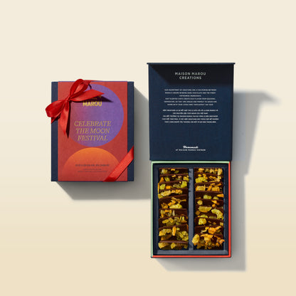 A gourmet chocolate gift set by Maison Marou Creations, featuring a dark navy blue box with a red ribbon on the left, and the open box on the right showing an assortment of chocolate bars with dried citrus slices inside.