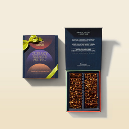 An open dark blue gift box labeled Maison Marou Creations and 'Celebrate the Moon Festival' is shown beside its matching closed outer box. The open box contains multiple compartments filled with assorted chocolates and nuts, photographed on a warm cream background with soft shadows.